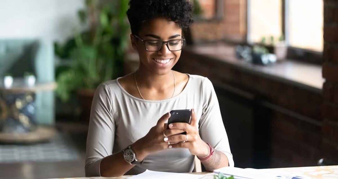 What if I Can’t Afford My Eczema Medications? 1 A smiling person with glasses sits at a table, looking at a smartphone. They are wearing a gray top and are in a well-lit room with plants and a large window in the background. Papers are scattered on the table.