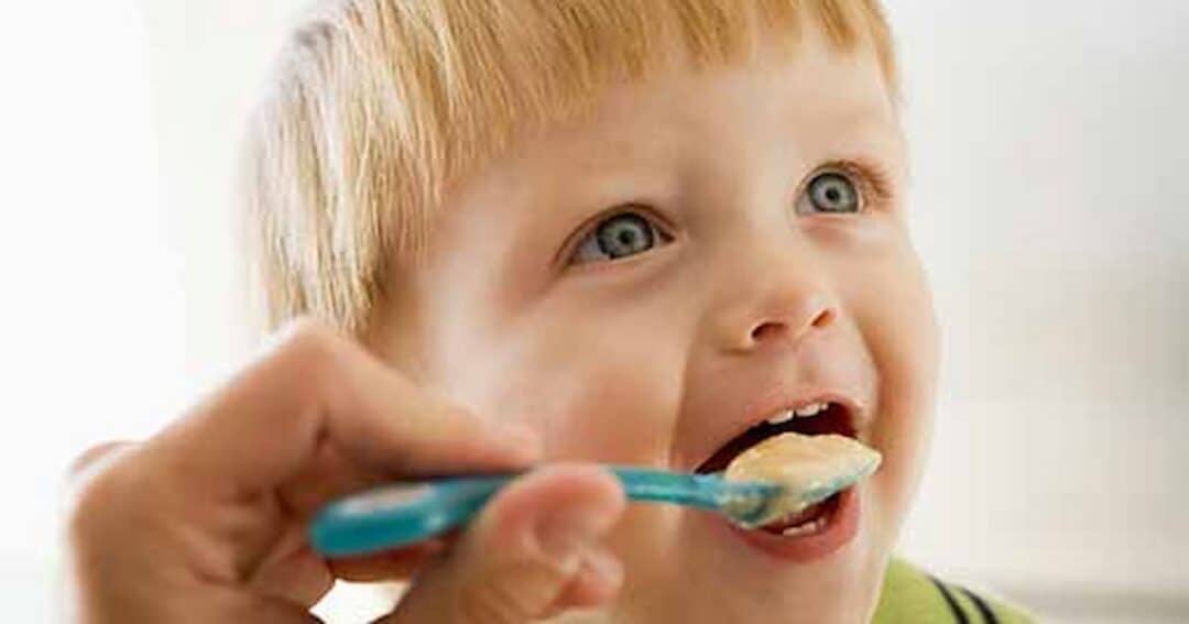 Food Allergies 5 A young child with blonde hair is being fed with a blue spoon. The child looks happy and eager, with an open mouth ready to eat the food from the spoon. The background is softly blurred.