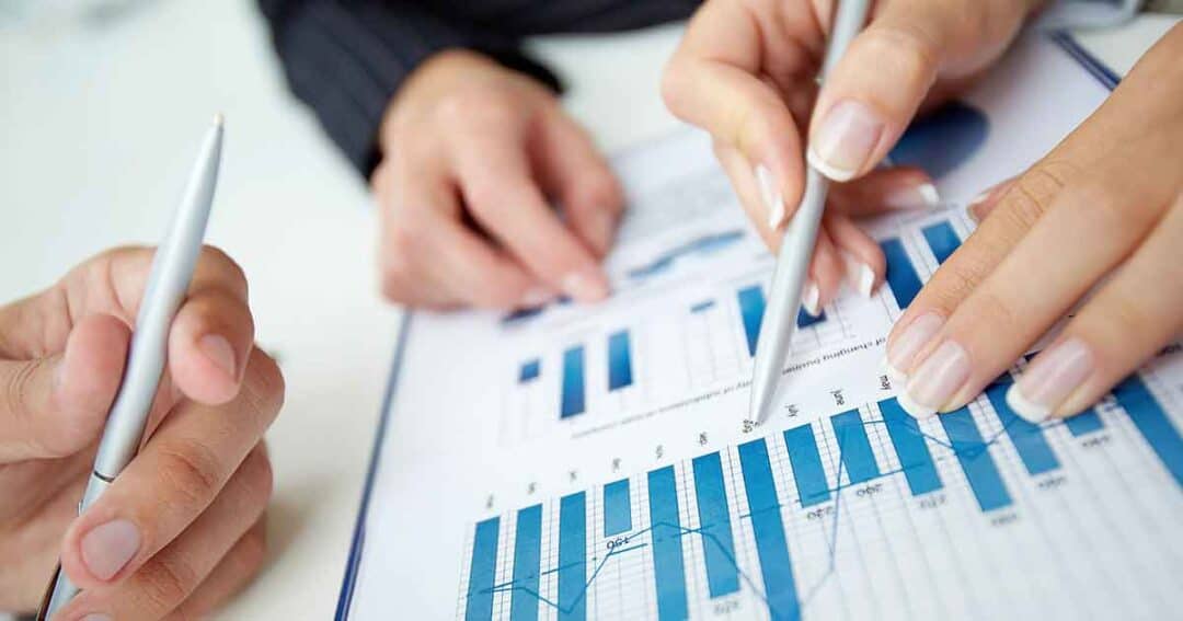 Close-up of two people discussing a financial chart. One person holds a pen pointing at a bar graph with blue bars, while the other holds a pen nearby. The chart includes various data points and lines on a clipboard.