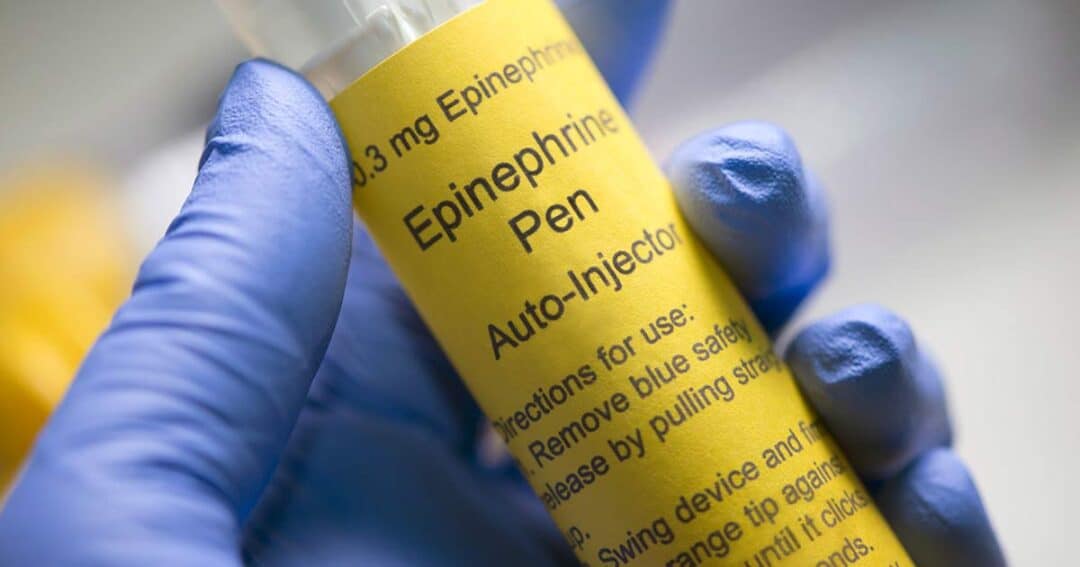 Anaphylaxis 9 Close-up of a gloved hand holding an EpiPen autoinjector with a yellow label. The label includes the words "0.3 mg Epinephrine Pen" and "Auto-Injector" along with usage instructions.