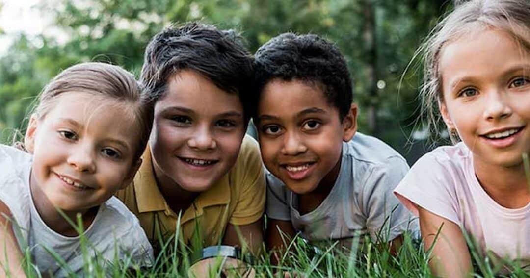 Four children are lying on the grass, smiling at the camera. The scene is set outdoors with greenery in the background. They appear happy and relaxed, enjoying a moment together in nature.