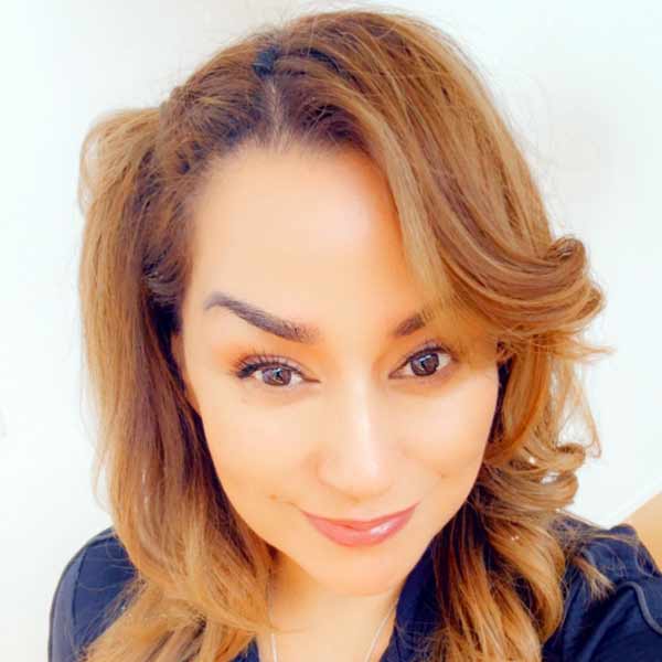 Unidos Hablemos Virtual Conference Series 4 A woman with wavy, light brown hair and subtle makeup smiles softly at the camera. She is wearing a dark top and is positioned against a plain light background.