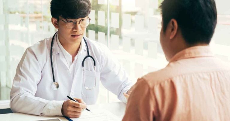 A doctor with glasses and a stethoscope around his neck sits at a desk, holding a pen and talking to a patient who is seated opposite him. The setting is a bright office with frosted windows in the background.