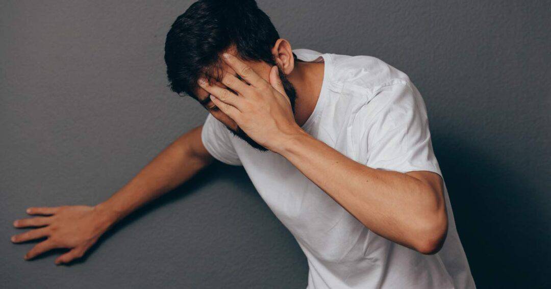 Anaphylaxis 3 A man in a white shirt leans against a gray wall, covering his face with one hand, as if warding off the onset of anaphylaxis. His expression reveals distress or headache, underscoring the urgency of his situation.