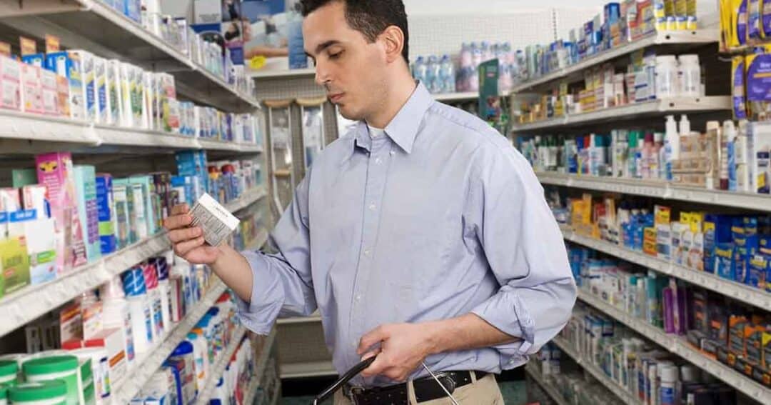 A man in a blue shirt is standing in a pharmacy aisle, holding a product and reading its label. Shelves stocked with various health products surround him. He is carrying a shopping basket.