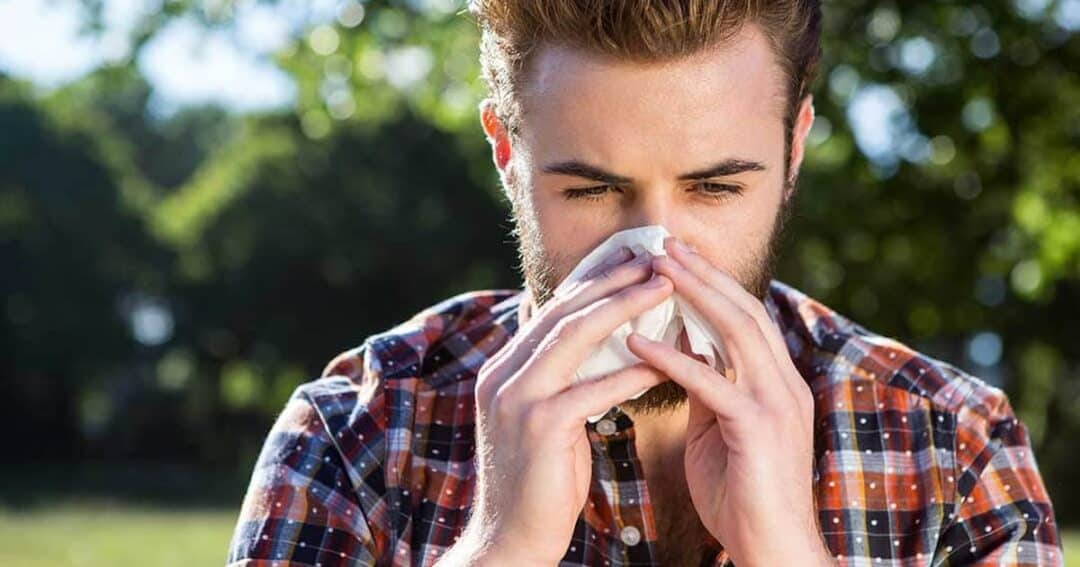 A man with a beard wearing a plaid shirt stands outdoors, holding a tissue to his nose as if sneezing or blowing his nose. Behind him are blurred trees and greenery, suggesting he is in a park or natural setting.