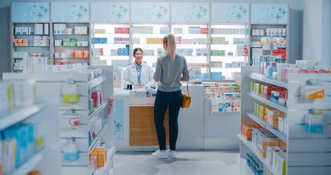 A woman stands at the counter of a pharmacy, speaking with a pharmacist. Shelves filled with various medications and health products are visible in the background. The space is brightly lit and organized.