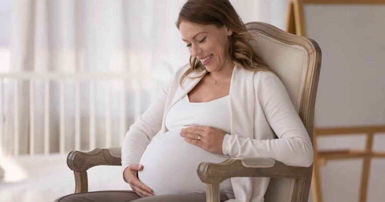 A pregnant woman sits in a cushioned chair, gently cradling her belly with both hands. She is smiling and looking down, wearing a white top and a light-colored cardigan. The background is softly lit and blurred, suggesting a calm, serene setting.