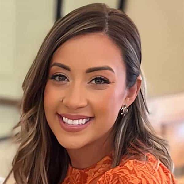 Unidos Hablemos Virtual Conference Series 2 Smiling woman with long, light brown hair wearing an orange patterned top, posing indoors.