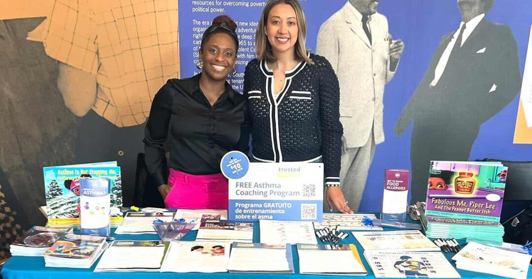 Two women stand behind a table with pamphlets and booklets about asthma coaching and health resources. A sign reads “Free Asthma Coaching Program.” The background features images of historical figures.