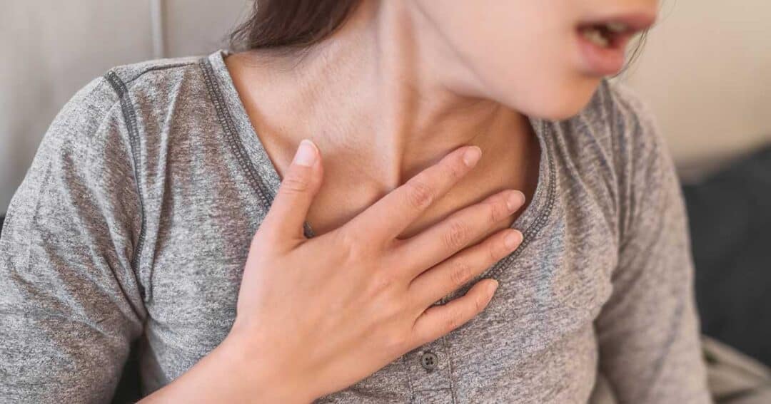 Anaphylaxis 1 A person wearing a gray top is clutching their chest, seemingly in discomfort or experiencing difficulty breathing, possibly due to anaphylaxis. Their face is partly visible, and their posture suggests concern.