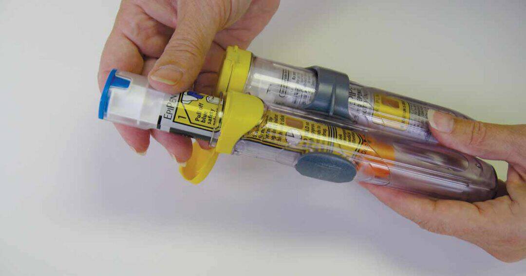 Anaphylaxis 5 Two hands hold two epipens, one with a yellow cap and the other with an orange cap, against a white background. The auto-injectors have labels and instructions printed on them.