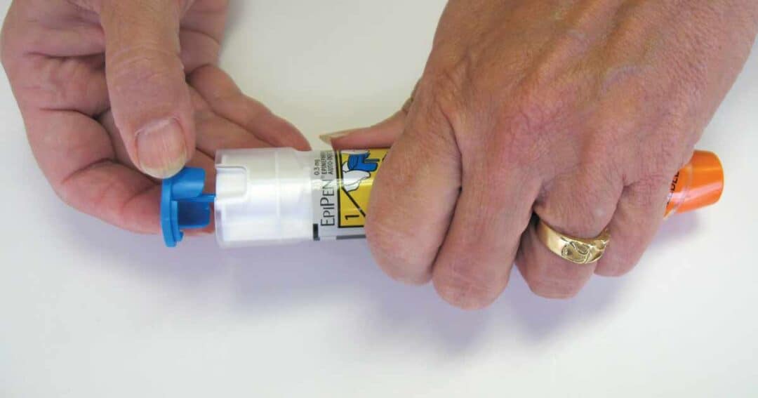 Anaphylaxis 10 A person carefully holds an EpiPen, their hands moving deftly to remove the blue safety cap. The device features a distinct yellow label and an orange tip, while a ring subtly adorns their left finger, reflecting light.