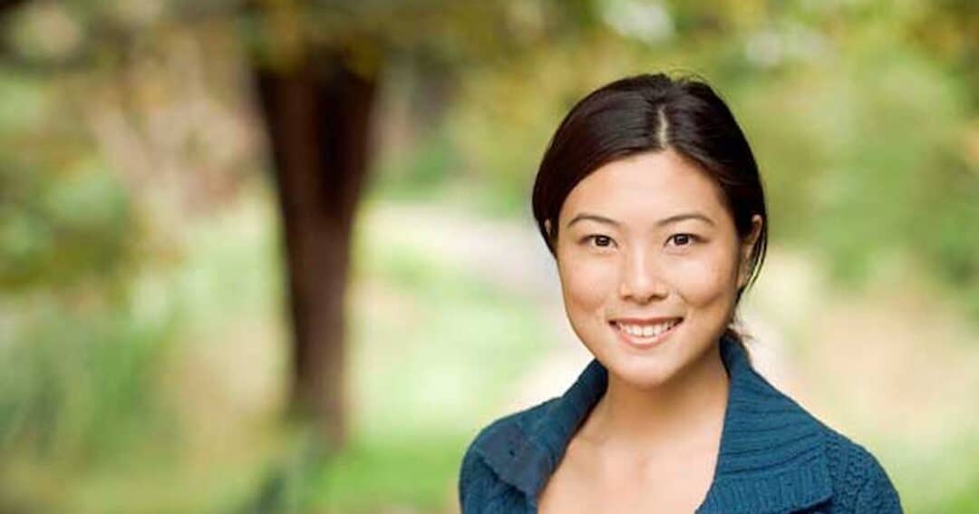 Allergy Diagnosis and Testing 1 A person with long hair smiles at the camera while standing outdoors. The background is a blurry, lush scene with green trees and grass. The individual is wearing a blue top.