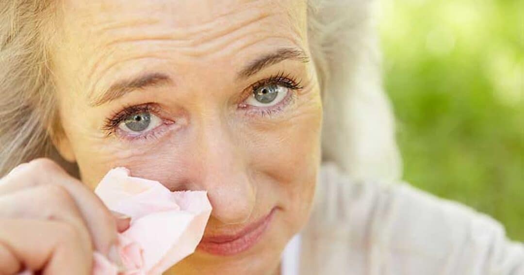 A woman with light-colored hair wipes her eye with a tissue while outdoors. She appears emotional or teary-eyed, with a background of greenery.